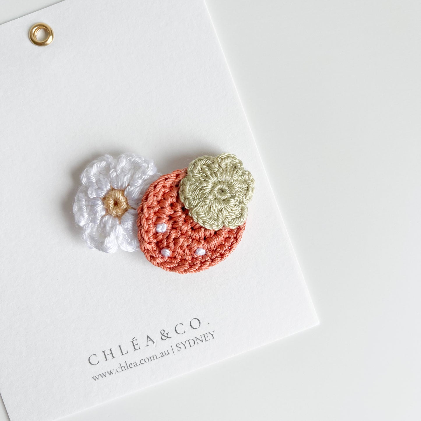 Handmade crochet clip of three small, handmade crochet appliqués (white flower, reddish-orange strawberry shape, and green flower) from CHLÉA & CO.